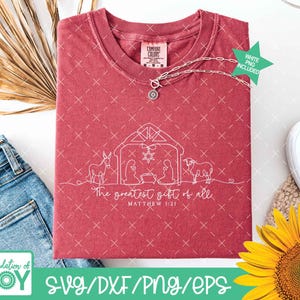 May include: A maroon Comfort Colors t-shirt featuring a white line drawing of a nativity scene with the text "The greatest gift of all" and "Matthew 1:21". A silver necklace with a pendant is draped across the shirt.