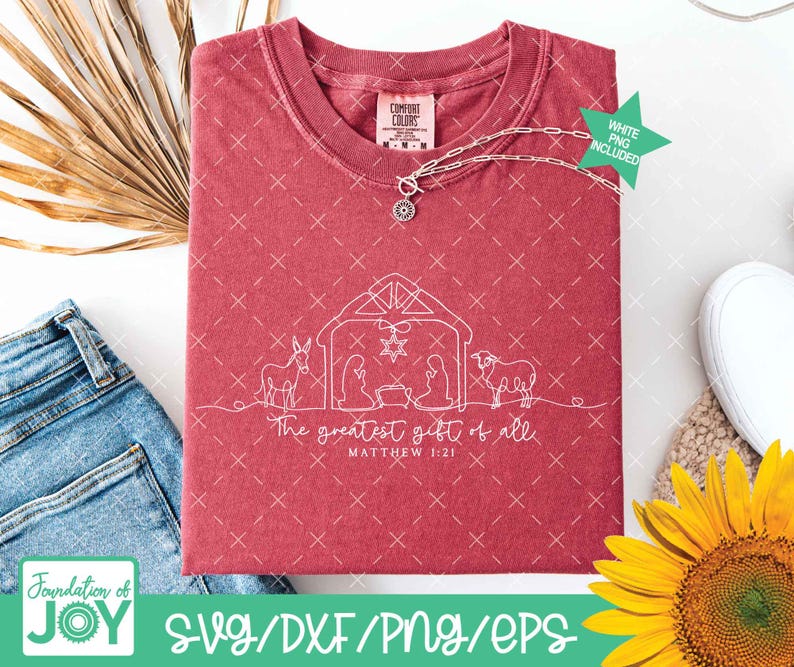 May include: A maroon Comfort Colors t-shirt featuring a white line drawing of a nativity scene with the text "The greatest gift of all" and "Matthew 1:21". A silver necklace with a pendant is draped across the shirt.