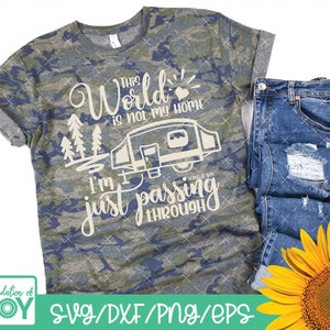 May include: A camouflage t-shirt with the text "This world is not my home I'm just passing through" and a small camper van graphic. The text is in a cream color. The t-shirt is on a white background with a pair of blue jeans and a sunflower.