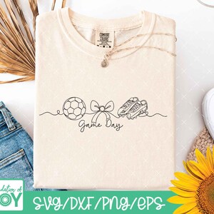 May include: Cream-coloured t-shirt featuring a black line drawing of a football, bow, and football boots, with the words "Game Day." A gold necklace and a Comfort Colors tag are visible. The shirt is next to blue jeans and a sunflower.