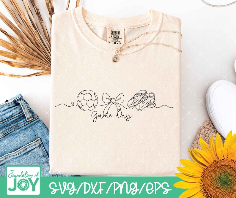 May include: Cream-coloured t-shirt featuring a black line drawing of a football, bow, and football boots, with the words "Game Day." A gold necklace and a Comfort Colors tag are visible. The shirt is next to blue jeans and a sunflower.