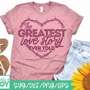 Pu&ograve; includere: Una maglietta rosa con un design a forma di cuore con un bordo di filo spinato. Il testo "The Greatest Love Story Ever Told" &egrave; all'interno del cuore con il riferimento biblico "1 John 4:10" sotto.