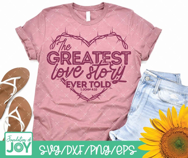 Pu&ograve; includere: Una maglietta rosa con un design a forma di cuore con un bordo di filo spinato. Il testo "The Greatest Love Story Ever Told" &egrave; all'interno del cuore con il riferimento biblico "1 John 4:10" sotto.