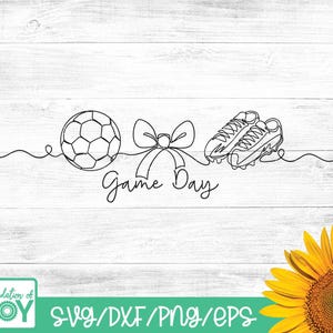 May include: Black and white line art design featuring a soccer ball, a bow, and soccer cleats. The words "Game Day" are written in cursive. The image is on a white wood background. A sunflower is in the bottom right corner.