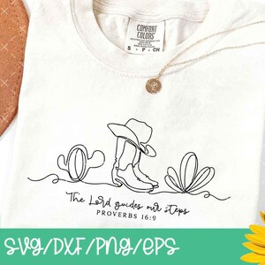 K&ouml;nnte beinhalten: Cremefarbenes T-Shirt mit einer schwarzen Strichzeichnung eines Cowboyhuts, Stiefel und Kakteen. Der Text "The Lord guides our steps" steht unter der Zeichnung. Eine goldene Halskette und ein brauner Hut sind ebenfalls zu sehen.