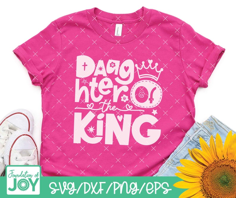 May include: Pink t-shirt with a white design that says "Daughter of the King" with a crown and a heart. The shirt has a white cross and a star.