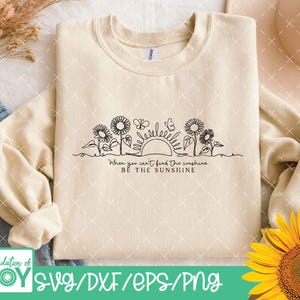 May include: Cream-colored sweatshirt with a black line drawing of sunflowers, butterflies, and a sun. The text "When you can't find the sunshine, be the sunshine" is below the drawing. The bottom of the image has the text "SVG/DXF/EPS/PNG".