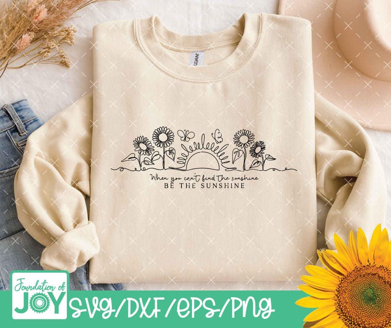 May include: Cream-colored sweatshirt with a black line drawing of sunflowers, butterflies, and a sun. The text "When you can't find the sunshine, be the sunshine" is below the drawing. The bottom of the image has the text "SVG/DXF/EPS/PNG".