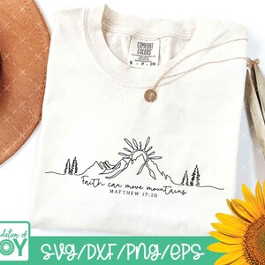May include: Cream-colored t-shirt with a black line drawing of mountains, sun, and trees. The text "Faith can move mountains Matthew 17:20" is below the drawing. A gold necklace and a sunflower are also in the image.