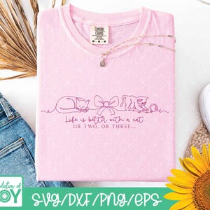 May include: A light pink Comfort Colors t-shirt with the text "Life is better with a cat or two or three." The shirt features a line drawing of two cats and a bow. A silver necklace with a flower pendant is draped on the shirt.