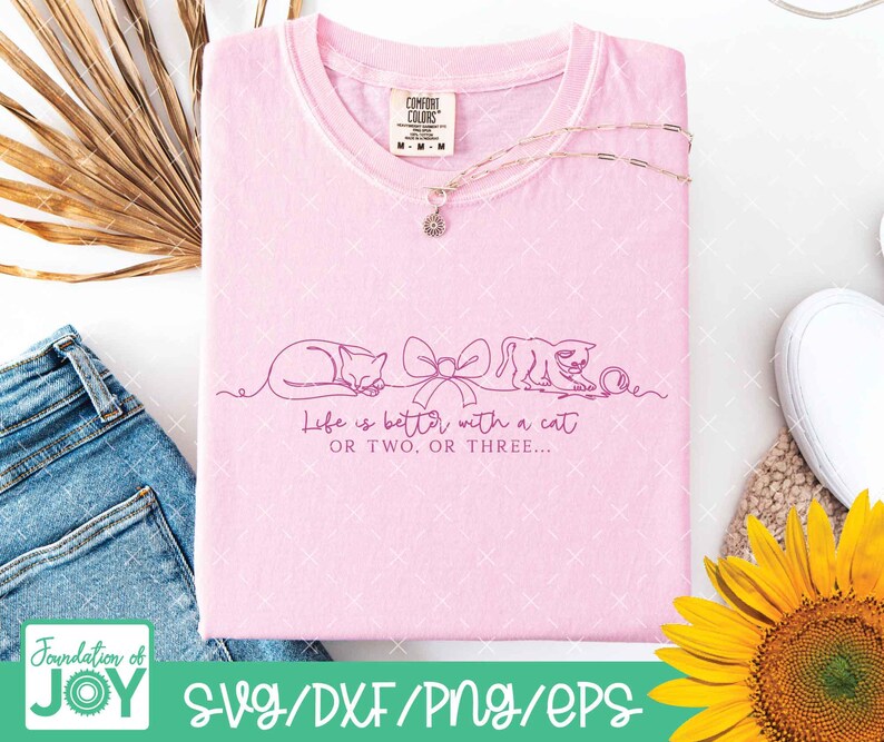 May include: A light pink Comfort Colors t-shirt with the text "Life is better with a cat or two or three." The shirt features a line drawing of two cats and a bow. A silver necklace with a flower pendant is draped on the shirt.