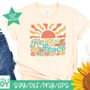 May include: A beige t-shirt with a retro-style graphic design featuring a sun, flowers, and the text "He will be our peace".