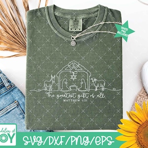 May include: An olive green t-shirt with a white line drawing of a nativity scene, including a star, animals, and figures. The text "The greatest gift of all" and "Matthew 1:21" are below the image. A silver necklace is draped across the shirt.