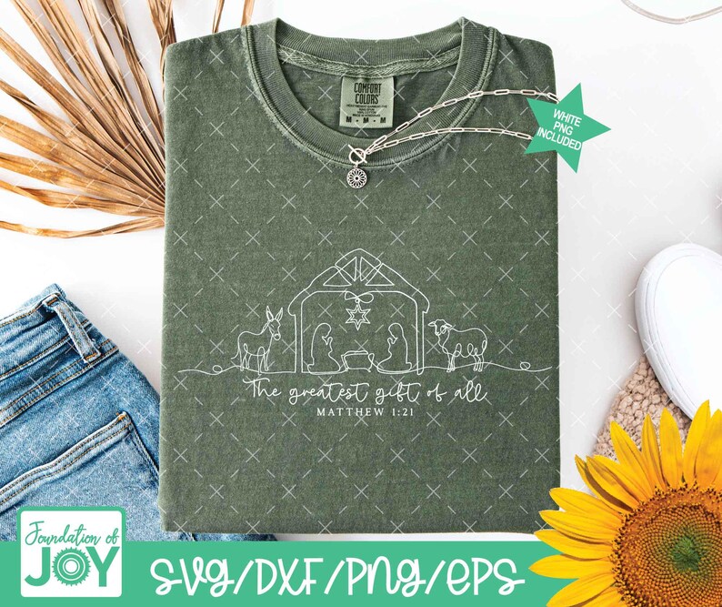 May include: An olive green t-shirt with a white line drawing of a nativity scene, including a star, animals, and figures. The text "The greatest gift of all" and "Matthew 1:21" are below the image. A silver necklace is draped across the shirt.