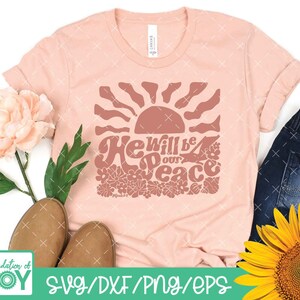 May include: A peach-colored t-shirt with a graphic design of a sun with rays and flowers. The text "He will be our peace" is written in a retro font style.