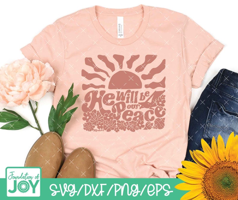 May include: A peach-colored t-shirt with a graphic design of a sun with rays and flowers. The text "He will be our peace" is written in a retro font style.