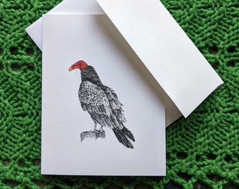 May include: A blank greeting card with a black and white illustration of a turkey vulture with a red head. The card is folded in half and has a white envelope.