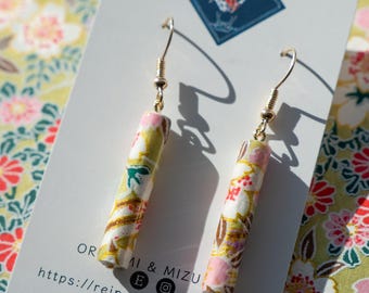 May include: Dangle earrings with gold-tone hooks and cylindrical charms. The charms are wrapped in patterned paper with pink, white, yellow, and gold floral designs. The earrings are displayed on a white card with text and a floral background.