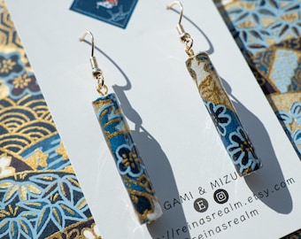 May include: Dangle earrings with a cylindrical design. The earrings have a blue and gold floral pattern and gold-coloured ear wires. The earrings are displayed on a white card with the text "GAMI & MIZU".