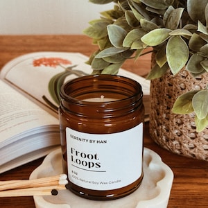 May include: A brown glass jar candle with a white label that reads "Froot Loops" and "100% Natural Soy Wax Candle." The candle sits on a white, scalloped tray with matches. A book and greenery are in the background.