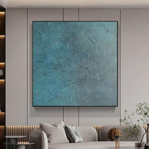 May include: A large, square abstract painting with a textured surface. The artwork features shades of blue and gray, with a black frame. The painting is displayed above a gray sofa in a modern living room setting.