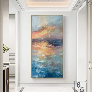 Op de afbeelding: Abstracte schilderij van een zeegezicht met een gouden zonsondergang die in het water wordt weerspiegeld. Het schilderij is ingelijst in een gouden lijst en hangt aan een witte muur.