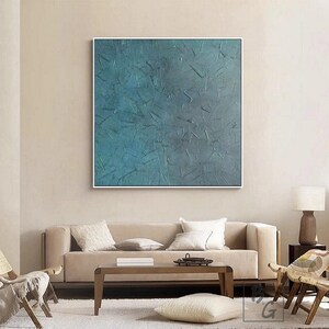May include: Large square abstract painting with a textured teal and blue surface, framed in white. The artwork hangs above a beige sofa with white pillows, a wooden coffee table, and a cream-colored rug. A lamp and chairs complete the living room setting.