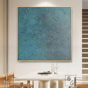 May include: Square abstract painting with a textured teal and blue surface, framed in gold. The artwork is displayed above a white marble table with chairs. The painting's surface has visible brushstrokes and a layered effect.
