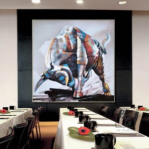 Puede incluir: Una pintura abstracta colorida de un toro cargando, con un esquema de colores blanco, azul, rojo y amarillo. El toro est&aacute; representado en una pose din&aacute;mica, con la cabeza baja y los cuernos apuntando hacia adelante. La pintura est&aacute; enmarcada en un marco negro y cuelga de una pared en una habitaci&oacute;n con una mesa y sillas blancas.