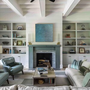 May include: A living room with a fireplace, built-in bookshelves, and a light-colored sofa and chairs. The fireplace is painted a muted blue-green, and a blue abstract painting hangs above it. The room has a neutral color palette.