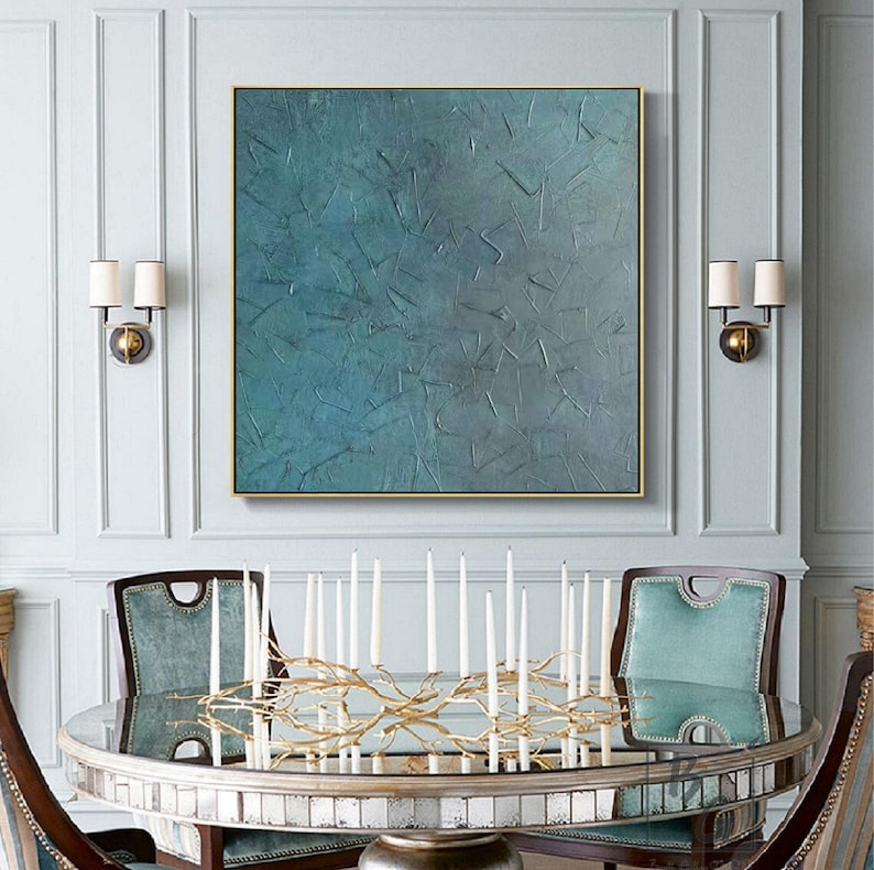 May include: An abstract teal painting in a gold frame hangs above a round mirrored table. The table is set with a gold branch-like candelabra holding white candles. Teal upholstered chairs surround the table, creating an elegant dining room setting.