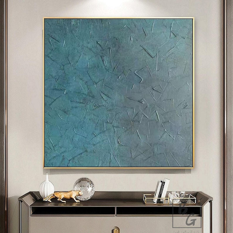 May include: Square abstract painting with a textured teal and blue surface, framed in gold. The artwork features a layered effect with visible brushstrokes, creating a dynamic visual texture. The painting is displayed above a dark cabinet with decorative items.