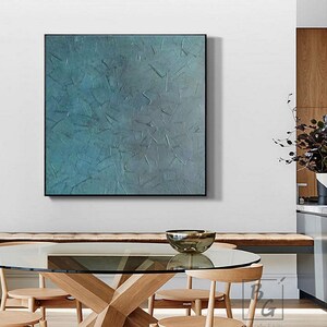 May include: Abstract teal and gray textured wall art in a black frame. The artwork features a layered, tactile surface with a variety of shapes and textures. The painting is displayed above a wooden table with a glass top and wooden chairs.