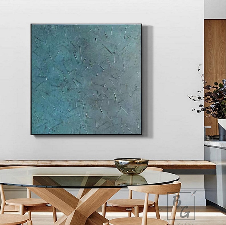 May include: Abstract teal and gray textured wall art in a black frame. The artwork features a layered, tactile surface with a variety of shapes and textures. The painting is displayed above a wooden table with a glass top and wooden chairs.