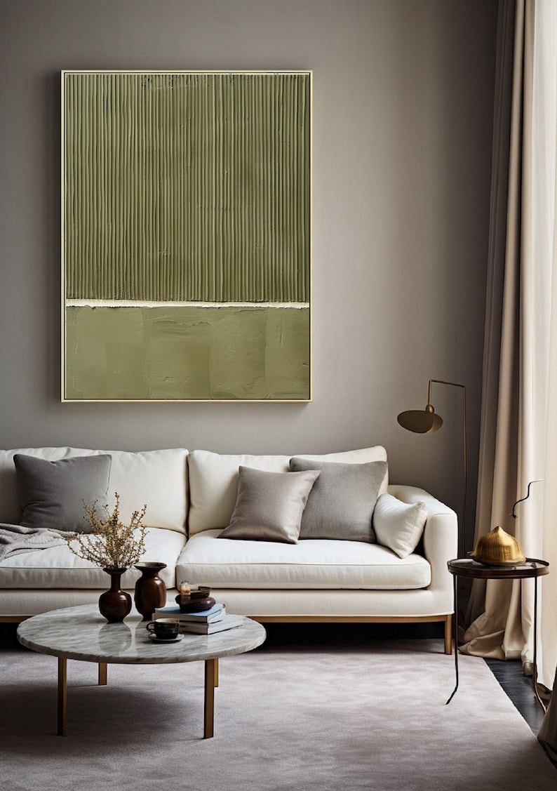 Op de afbeelding: Een witte bank met twee grijze kussens staat voor een groen-wit abstract schilderij. Een kleine ronde marmeren salontafel met een vaas met gedroogde bloemen en boeken staat voor de bank. Een gouden staande lamp en een bijzettafel met een gouden theepot bevinden zich op de achtergrond.