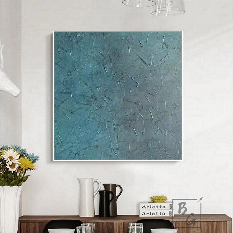 May include: Square abstract painting with a textured teal and blue surface. The artwork is framed in a white border and hangs on a white wall. The painting has a modern, contemporary style.