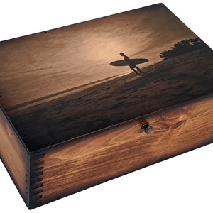 May include: A wooden box with a dark brown stain and a silhouette of a person surfing on the beach on the lid. The box has a brass latch.