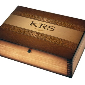May include: A wooden box with a brown stain and a gold-colored top. The top has a decorative border and the initials "KRS" are engraved in the center.