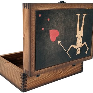 Puede incluir: Una caja de madera con una tapa con bisagras. La tapa presenta un fondo negro con un coraz&oacute;n rojo, tres puntos rojos y una silueta blanca de una persona colgada boca abajo sosteniendo una lanza. La caja est&aacute; hecha de madera marr&oacute;n oscura.