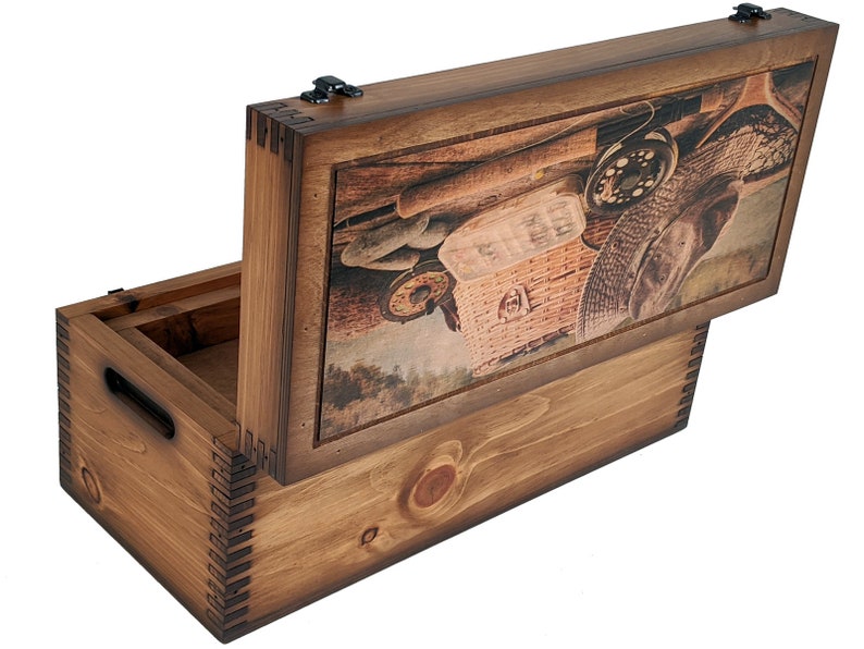 May include: A wooden box with a hinged lid. The lid features a printed image of fishing gear, including a fishing rod, a fishing reel, a hat, and a wicker basket. The box is stained a dark brown color.