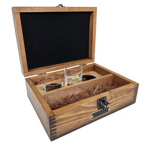 Puede incluir: Una caja de madera con tapa abatible, forrada de fieltro negro, y que contiene dos vasos de chupito de vidrio transparente. La caja tiene un cierre de lat&oacute;n y una peque&ntilde;a placa de nombre de lat&oacute;n.