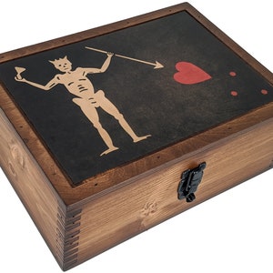 Puede incluir: Una caja de madera con una tapa con bisagras y un cierre de metal negro. La tapa presenta una bandera pirata negra y dorada con un dise&ntilde;o de calavera y huesos cruzados. La bandera tambi&eacute;n incluye un coraz&oacute;n rojo con una flecha que lo atraviesa y dos puntos rojos.
