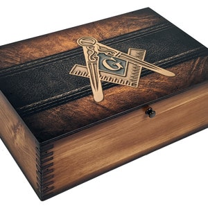 May include: A wooden box with a dark brown stain and a black leather-like band across the top. The box has a brass Masonic symbol on the lid. The symbol is a square and compass with a 'G' in the center.