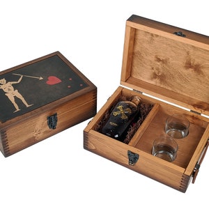 Puede incluir: Una caja de madera con tapa abatible, que contiene una botella de ron oscuro y dos vasos de chupito. La caja est&aacute; decorada con una bandera pirata con una calavera y huesos cruzados.
