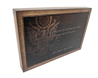Puede incluir: Placa de madera con una superficie oscura similar al cuero. El texto del juramento de la Constitución de los Estados Unidos está grabado en la superficie, junto con un emblema de águila. La placa está enmarcada en madera marrón claro.