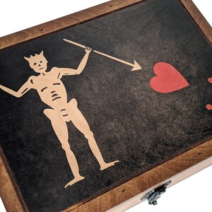 Puede incluir: Una caja de madera con tapa con bisagras y cierre de metal. La tapa presenta un fondo negro con un esqueleto blanco sosteniendo una lanza y un coraz&oacute;n rojo con dos puntos rojos.