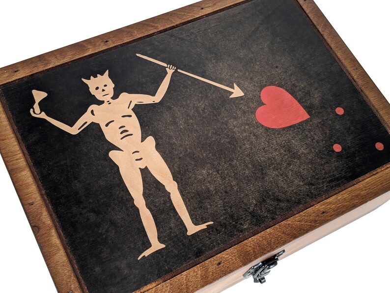 Puede incluir: Una caja de madera con tapa con bisagras y cierre de metal. La tapa presenta un fondo negro con un esqueleto blanco sosteniendo una lanza y un coraz&oacute;n rojo con dos puntos rojos.