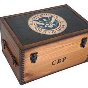 May include: A wooden box with a black lid featuring the U.S. Department of Homeland Security seal. The box has a brown stain and is engraved with "CBP".