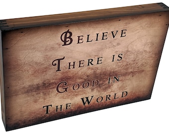 Könnte beinhalten: Ein Holzschild mit einem verwitterten braunen Hintergrund und schwarzen Buchstaben. Das Schild lautet "Glaube, dass es Gutes in der Welt gibt".