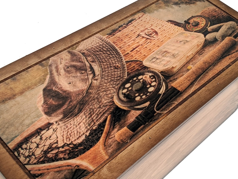 May include: A wooden box with a hinged lid. The lid features a printed image of fishing gear, including a hat, fishing rod, tackle box, and a fishing net. The background is a wood grain pattern.